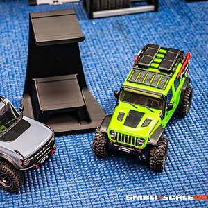 Double-decker display stand for 1/24 & 1/18 RC crawlers with two RC vehicles on blue surface.