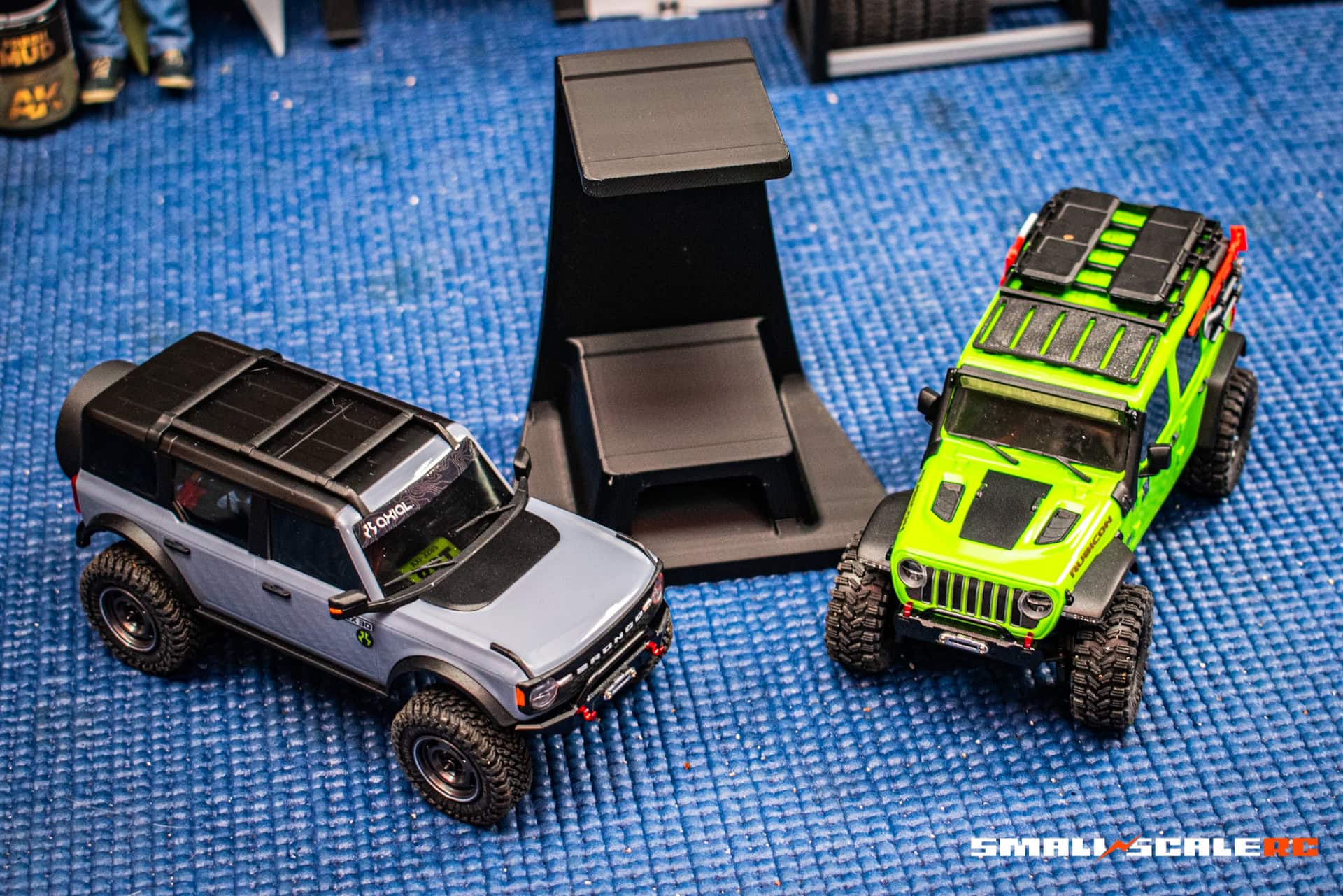 Double-decker display stand for 1/24 & 1/18 RC crawlers with two RC vehicles on blue surface.